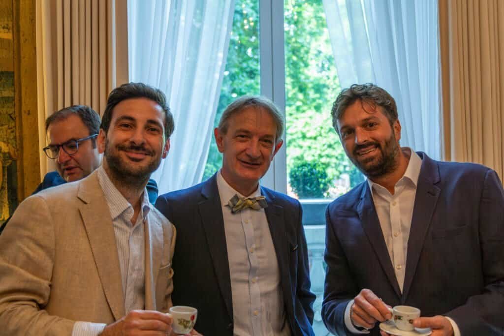 Group of three men smiling and holding teacups during a social event indoors.