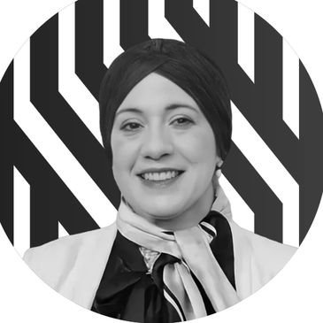 A portrait of a woman with short dark hair, smiling, wearing a light blazer and scarf, against a bol.