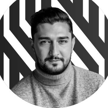 A confident young man with a beard and styled hair, wearing a sweater, in front of a geometric black.