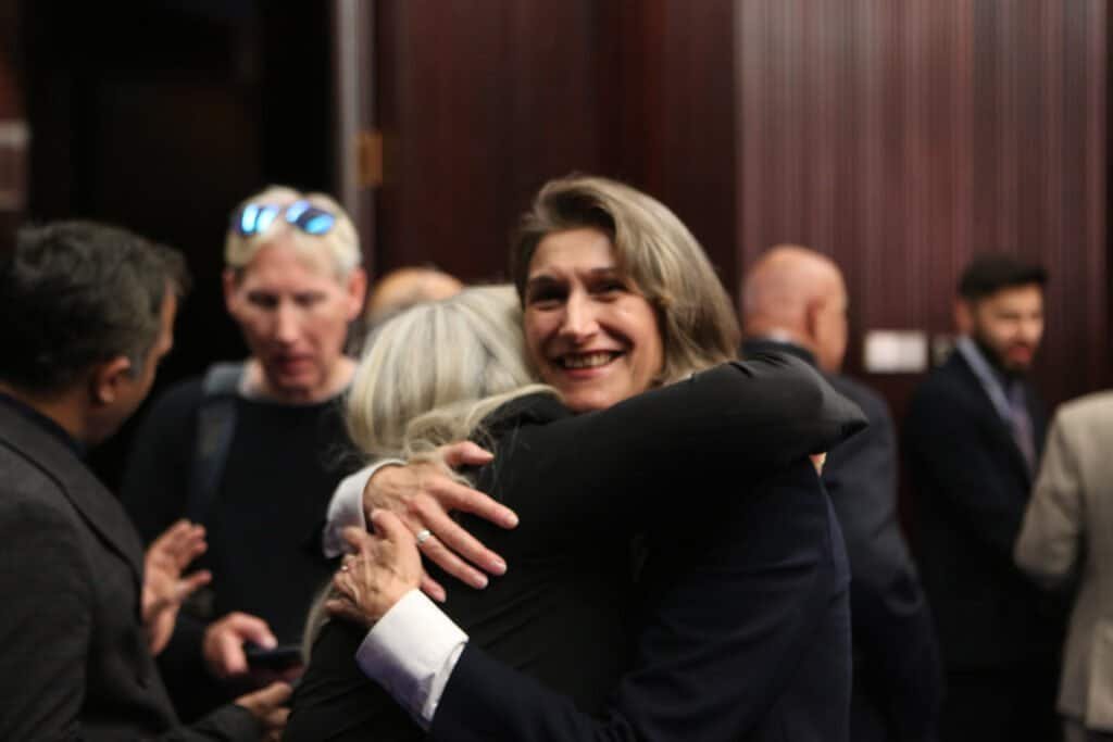 Two women hugging warmly at a professional event with others in the background.